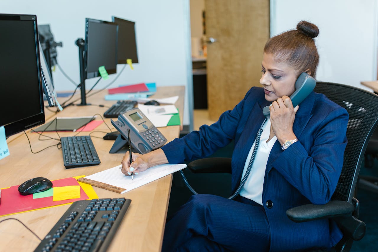 Businesswoman handling a phone call while writing notes, surrounded by computers and office supplies.