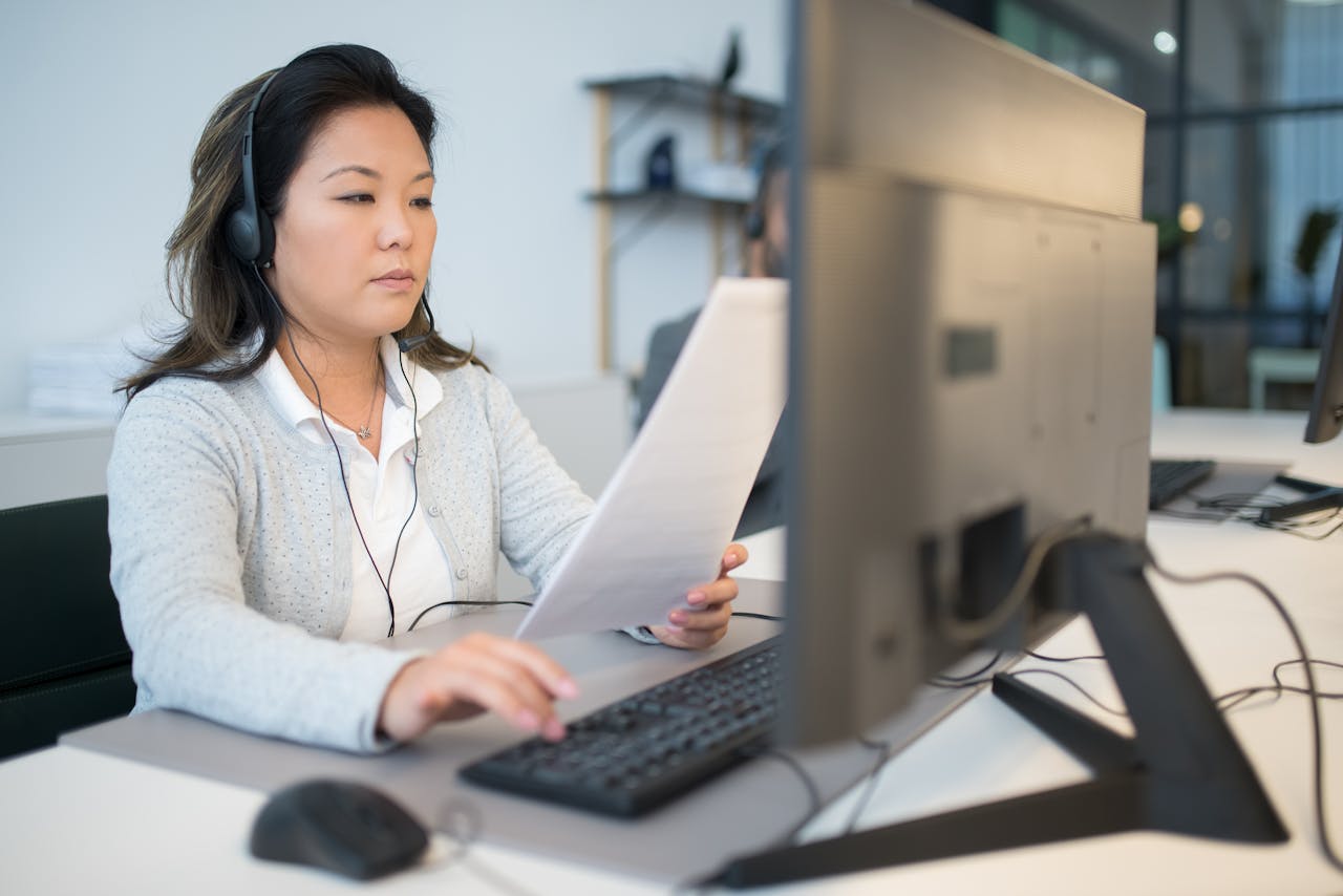 Professional woman wearing a headset and reading paper while working on a computer in an office setting.